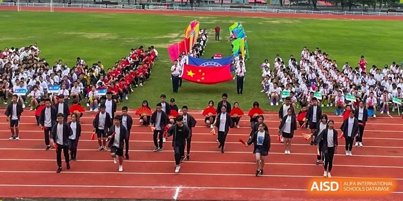Students from Wuhan Foreign Languages School International Division running on the track during a large parade or opening ceremony, led by a large Chinese flag.