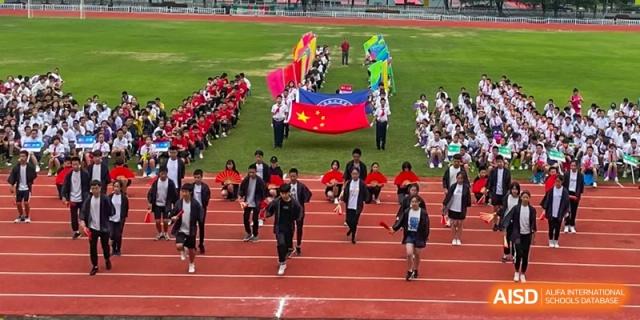 Students from Wuhan Foreign Languages School International Division running on the track during a large parade or opening ceremony, led by a large Chinese flag.