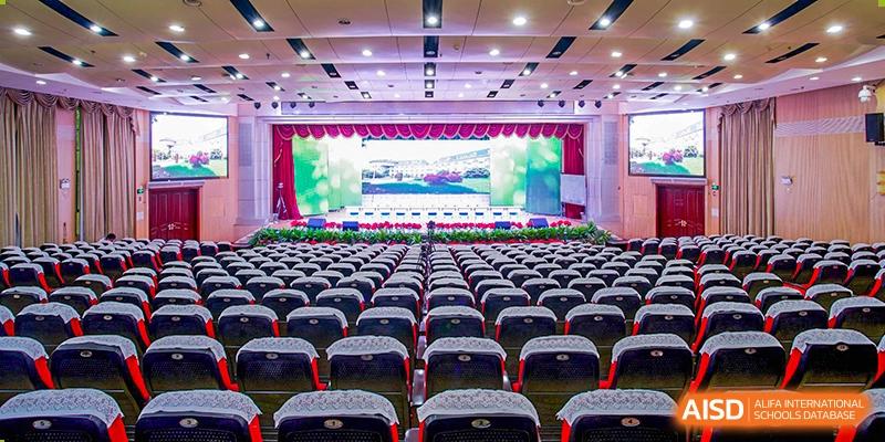A large school assembly or event in a modern hall with many rows of seating facing a brightly lit stage at Wuhan Foreign Languages School International Division.