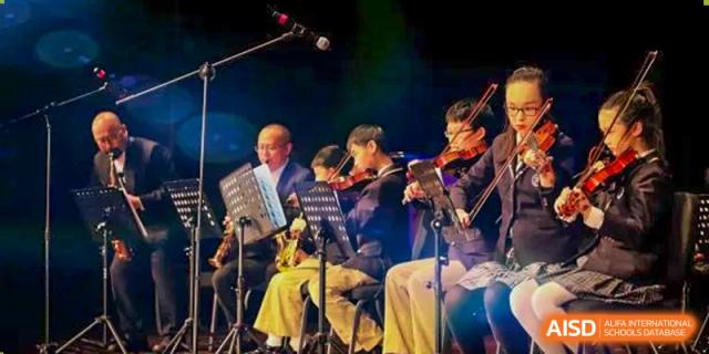 Students and teachers from Beijing Xin Fuxue International Academy perform together in an orchestra on a stage.