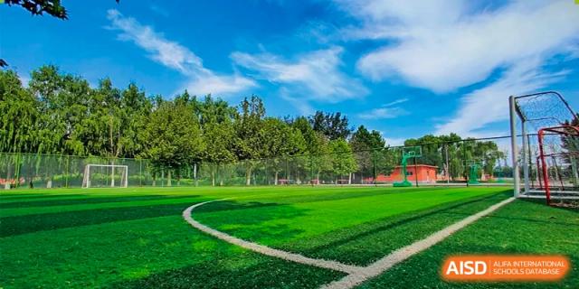 A wide, green sports field at Beijing Xin Fuxue International Academy with trees and buildings in the background.