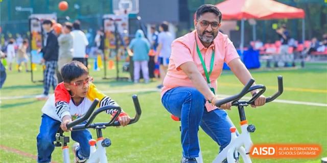 A man wearing a pink shirt and jeans smiles while sitting on an exercise bike at an event at Dulwich College Beijing.
