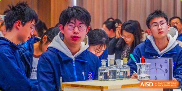 A close-up portrait of a male student in glasses and a blue jacket looking up from a science experiment at Wuxi Kuangyuan Bilingual School.