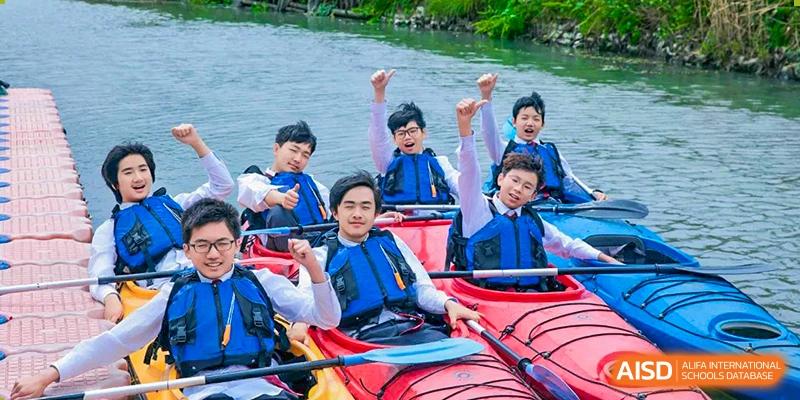 Students from Xi'an Jiaotong-Liverpool University Affiliated School (XJTLU-AS) are smiling and waving while in a red kayak on the water, wearing life vests.