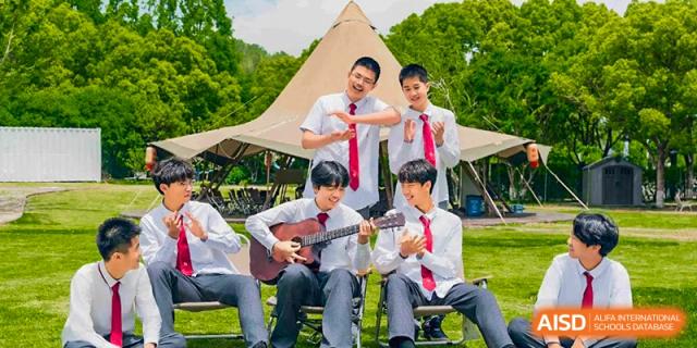 A group of male students from Xi'an Jiaotong-Liverpool University Affiliated School (XJTLU-AS) are sitting outdoors in front of a tent. One student is playing a guitar while the others pose and clap.