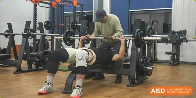 Two students from Xi'an Jiaotong-Liverpool University Affiliated School (XJTLU-AS) are in a weight room, one bench pressing with a spotter.