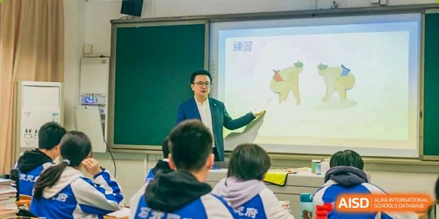 A teacher in a green jacket pointing to a map on a whiteboard while speaking to students in a classroom. Chengdu Shishi Tianfu High School International Department.