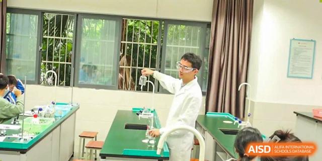 A male student in a white lab coat smiling while looking at a beaker in a science lab. Chengdu Shishi Tianfu High School International Department.
