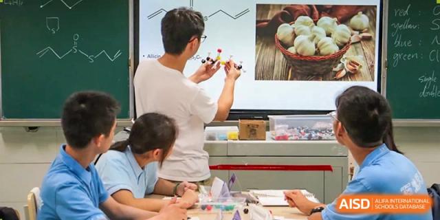 A male student pointing to a large screen that shows a molecular model, with other students and a teacher looking on. Chengdu Shishi Tianfu High School International Department.