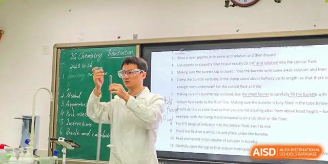A teacher in a lab coat and safety goggles holding up two beakers while giving a presentation in a classroom. Chengdu Shishi Tianfu High School International Department.