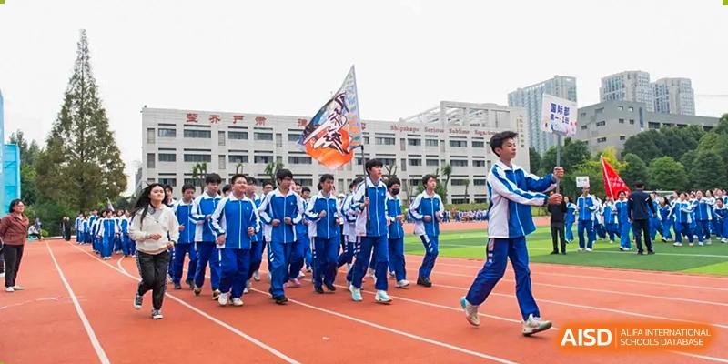 A group of students in blue tracksuits marching with a flag on a running track. Chengdu Shishi Tianfu High School International Department.