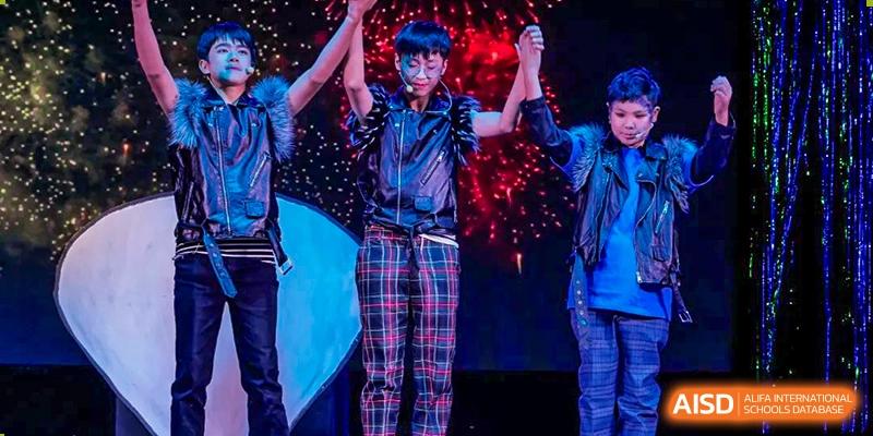 A student in plaid pants and sunglasses holding up their hands, possibly at a BaiShan AWS High School concert or talent show.