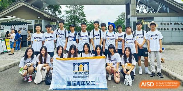 Students in matching white t-shirts posing with a banner for an international youth volunteer group at BaiShan AWS High School.