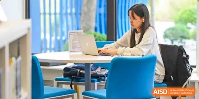 A student at Shanghai American School is using a laptop at a table with a blue chair.