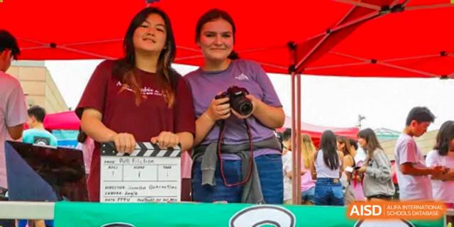 Two smiling female students at Shanghai American School are holding a camera and a film clapperboard at an event.