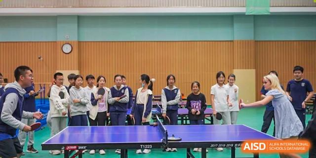 Group of students and adults standing behind a blue ping pong table in an indoor sports area, Beijing Etown Academy International Department