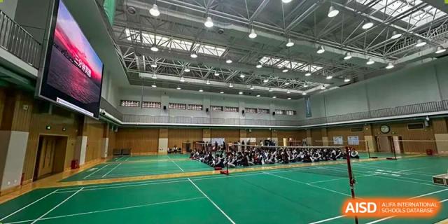 Wide view of a large, well-lit indoor gymnasium with an assembly of people on the green floor in the distance, Beijing Etown Academy International Department