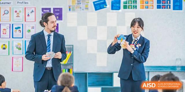 A teacher from Cogdel Cranleigh School Changsha standing and talking to a student seated at a desk, with a checkered wall design in the background.