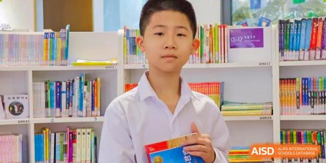 A male student from Cogdel Cranleigh School Changsha in a white polo shirt standing in the school library and holding a book.