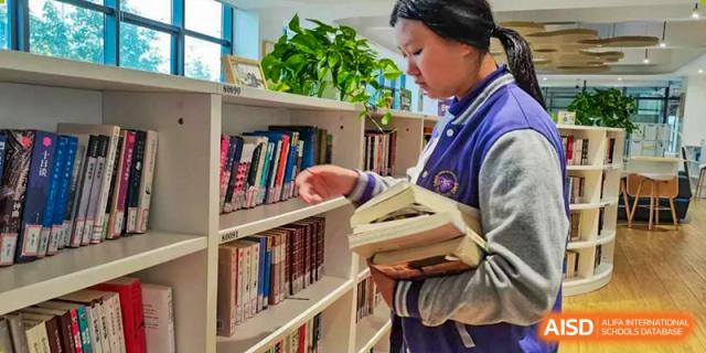A female student from Cogdel Cranleigh School Changsha browsing the shelves and holding books in the school library.