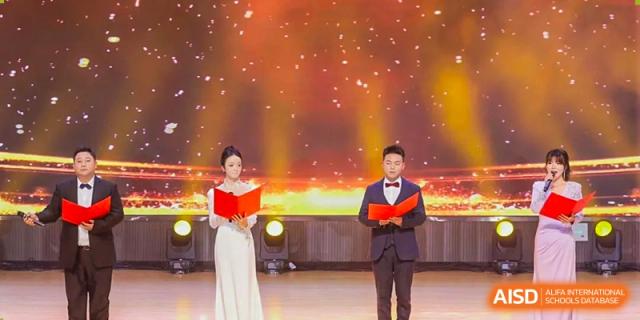 A male and female host or speaker from Cogdel Cranleigh School Changsha standing on a stage during a formal school event, both holding red books.