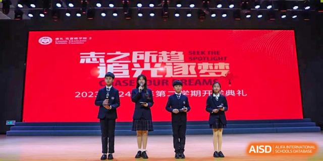 Four students from Cogdel Cranleigh School Changsha standing on a stage in uniform as hosts or speakers for a school event with a large red screen backdrop.