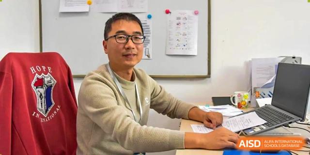 A male teacher or staff member at AISB-Hope International sitting at a desk and smiling, with a whiteboard in the background.