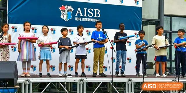 A group of young AISB-Hope International students performing on an outdoor stage with prop instruments.