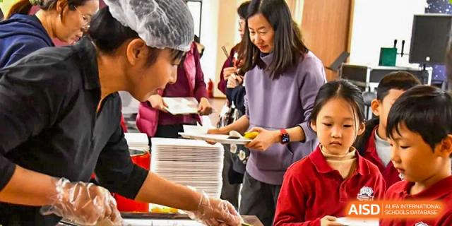 Staff members and a parent at AISB-Hope International serving food from a stack of plates at a school event or cafeteria.