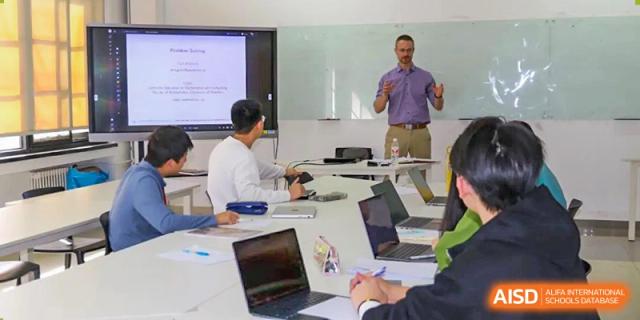A teacher from The Affiliated High School of Peking University's Dalton Academy giving a presentation or lecture to a class of students who are taking notes on laptops.
