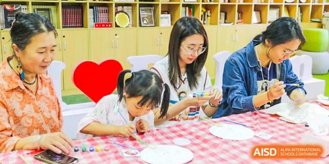 A teacher or older student from The Affiliated High School of Peking University's Dalton Academy assisting two younger children with a craft or writing activity at a small table.