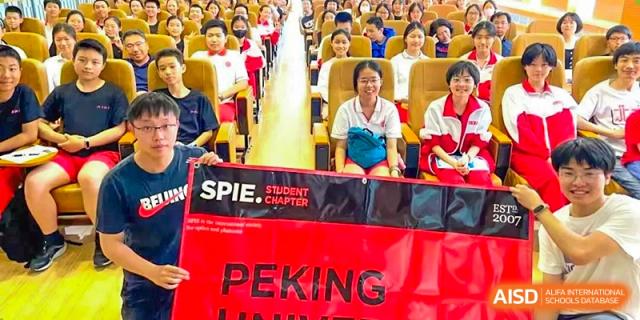 A student from The Affiliated High School of Peking University's Dalton Academy seated during an event or competition, with a red banner that reads "PEKING" prominently displayed.