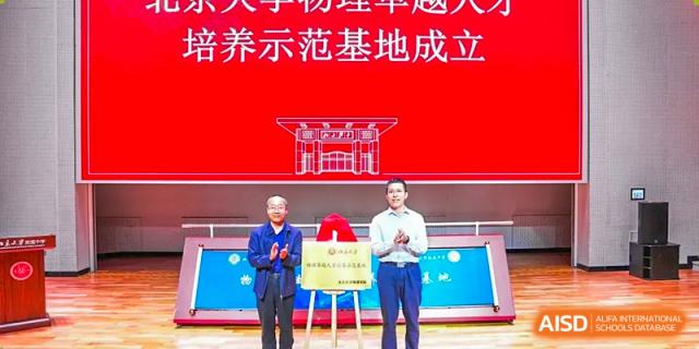 Two speakers, including one holding a plaque, on a stage during an event for The Affiliated High School of Peking University's Dalton Academy, beneath a large red banner.