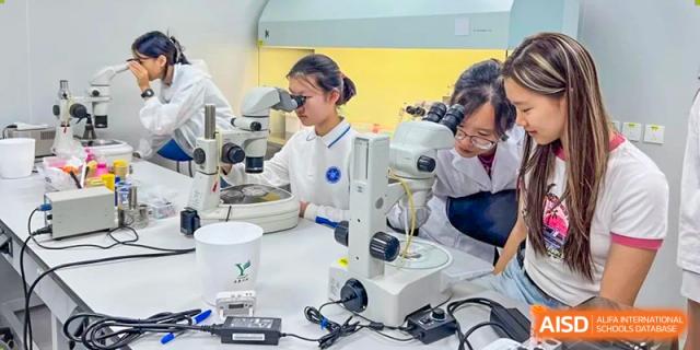 Students from The Affiliated High School of Peking University's Dalton Academy wearing lab coats and safety glasses while working simultaneously on different microscopes in a laboratory.