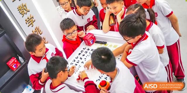 Young students from The Affiliated High School of Peking University's Dalton Academy wearing red-rimmed glasses and collaborating closely on a hands-on activity or puzzle at a table.