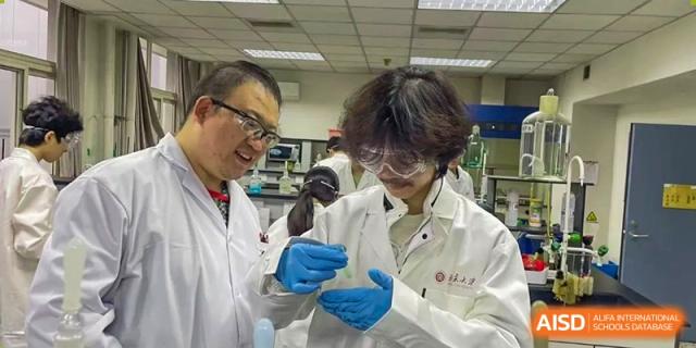 A teacher from The Affiliated High School of Peking University's Dalton Academy wearing a lab coat and safety glasses, smiling while instructing or assisting a student in the science lab.