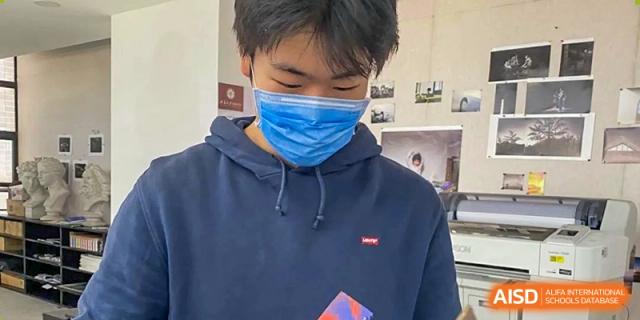A male student from The Affiliated High School of Peking University's Dalton Academy wearing a blue hoodie and a protective face mask, looking down intently at an object in his hands.