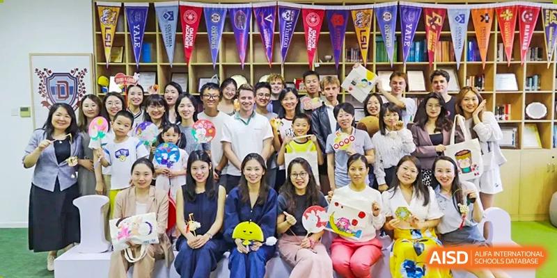 A large group of students and teachers posing for a cheerful group photo indoors, surrounded by colorful flags and decorations at The Affiliated High School of Peking University's Dalton Academy.