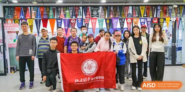 A group of students and chaperones from The Affiliated High School of Peking University's Dalton Academy posing for a photo while holding a red school banner, with various university pennants hanging in the background.