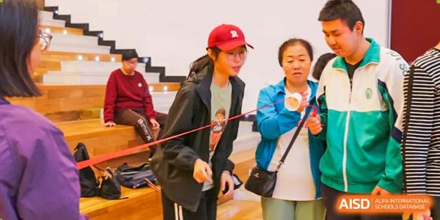 A student in a baseball cap and a woman from The Affiliated High School of Peking University's Dalton Academy focused on an indoor activity or game involving a small cup.
