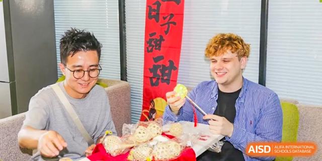 A foreign student from The Affiliated High School of Peking University's Dalton Academy smiling while holding a decorated paper lantern or food item, with a Chinese banner in the background.