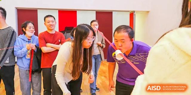 Students and a teacher from The Affiliated High School of Peking University's Dalton Academy participating in a fun indoor game, likely involving cups and string or straws.