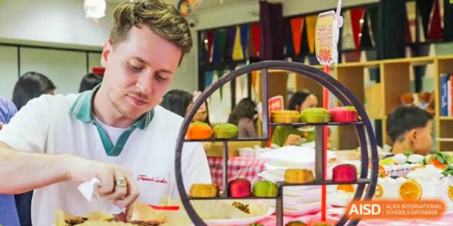 A teacher from The Affiliated High School of Peking University's Dalton Academy standing in front of a display of mooncakes and fresh fruit, likely at a cultural festival.