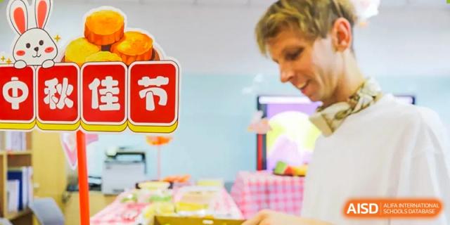 A close-up of a teacher from The Affiliated High School of Peking University's Dalton Academy smiling while handling food or supplies, with a large red Chinese character decoration in the foreground.
