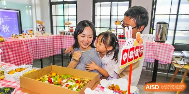 A woman and a young child from The Affiliated High School of Peking University's Dalton Academy happily looking at a box of colorful treats or fruit, with a sign featuring a rabbit icon.