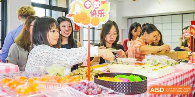 A group of teachers and students from The Affiliated High School of Peking University's Dalton Academy gathered around a table with a large platter of fruit and snacks, with decorations reading "Happy."
