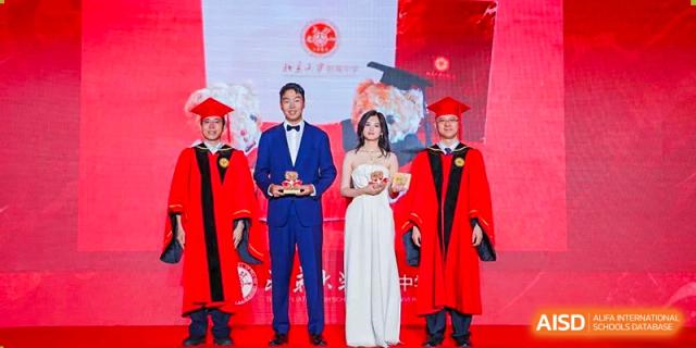 Two men in red graduation robes and a woman in a long white dress standing on a stage at an event for The Affiliated High School of Peking University's Dalton Academy, with a projected background.
