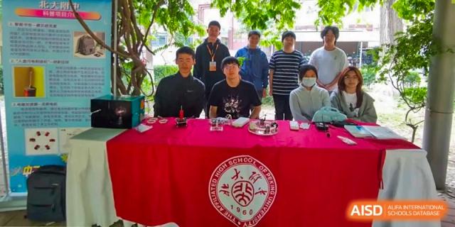A group of students, faculty, or staff from The Affiliated High School of Peking University's Dalton Academy sitting behind a red tablecloth with the school's logo, posing for a group photo.