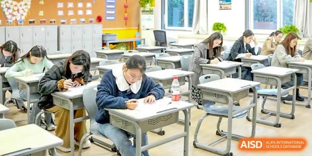 A focused CHONGQING WEIMING SCHOOL student writing or taking notes at a desk in an empty or sparsely occupied classroom.
