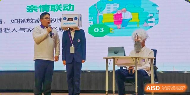 A student from Cogdel Cranleigh School Changsha in a suit and tie standing on a stage with a cardboard box costume head during a playful presentation.